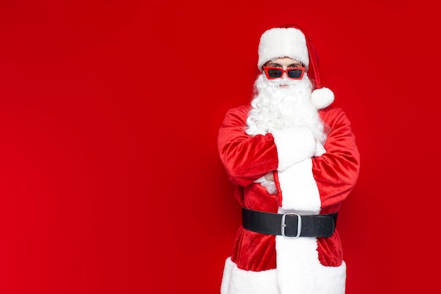 Ernsthafter Weihnachtsmann mit roter Brille steht mit verschränkten Armen auf farbigem Hintergrund