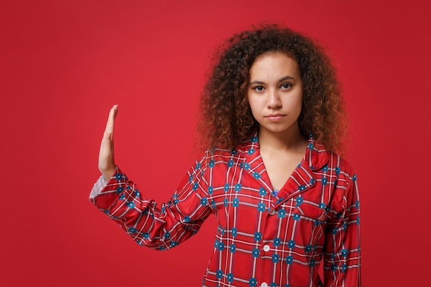 Ernsthaftes junges afrikanisch-amerikanisches Mädchen in Pyjama-Homewear, das zu Hause ruht, isoliert auf rotem Hintergrund. Entspannen Sie sich bei guter Laune Lifestyle-Konzept. Mock-up-Kopienbereich. Stop-Geste mit der Handfläche zur Seite zeigen.