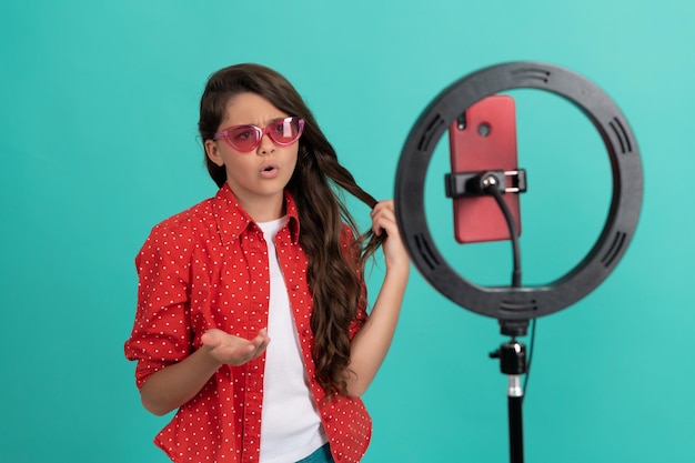 Ernsthaftes Kind mit Sonnenbrille, das vor der Kamera spricht, zeigt sein langes Haar, das online mit Selfie-LED bloggt, Kindheit.