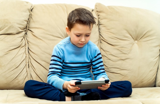 Ernsthaftes Kind sitzt mit seinen Beinen auf einem Sofa und spielt zu Hause auf dem elektronischen Gerät Kleiner Junge im blauen Pullover sitzt auf einer weichen Couch und spielt auf einem Tablet