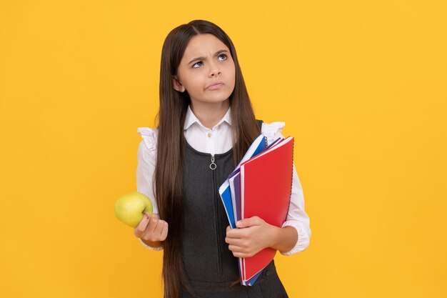 Ernsthaftes Mädchen im Schulalter denken, Apfel und Bücher halten gelbes Hintergrunddenken