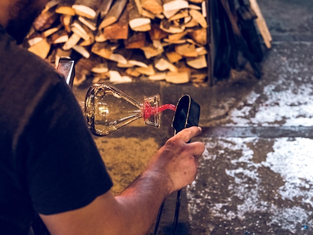 Ernten Sie einen nicht wiederzuerkennenden männlichen Arbeiter, der Glasvasen herstellt, während er in einer professionellen Werkstatt mit Brennholz bei Tageslicht arbeitet