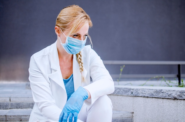 Foto erschöpfte ärztin sitzt vor der klinik und schaut weg. müde krankenschwester entspannt sich auf krankenhaustreppen. covid-19 virus world pandemie