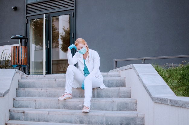 Erschöpfte Ärztin sitzt vor der Klinik und schaut weg. Müde Krankenschwester entspannt sich auf Krankenhaustreppen. Covid-19 Virus World Pandemie