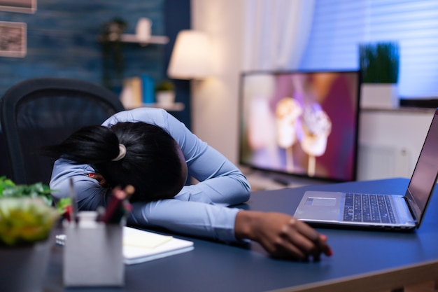 Erschöpfte dunkelhäutige Frau, die spät in der Nacht auf dem Schreibtisch im Heimbüro ruht. Beschäftigter Mitarbeiter, der modernes Technologienetzwerk verwendet, um Überstunden auf dem Tisch zu schlafen.
