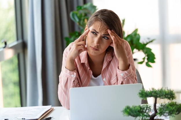 Erschöpfte Geschäftsfrau mit Kopfschmerzen im modernen Büro Kreative Frau, die am Schreibtisch arbeitet und sich müde fühlt Gestresste lässige Geschäftsfrau, die Kopfschmerzen bei der Überarbeitung des Desktop-Computers verspürt