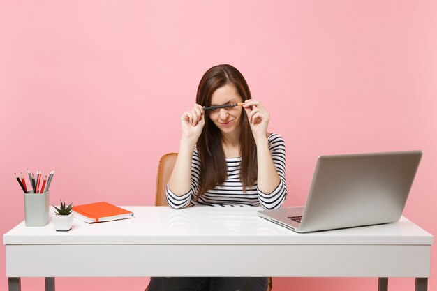 Erschöpfte müde Frau, die Probleme hat, den Bleistift in der Nähe des Gesichts zu halten, sitzt am weißen Schreibtisch mit einem modernen PC-Laptop