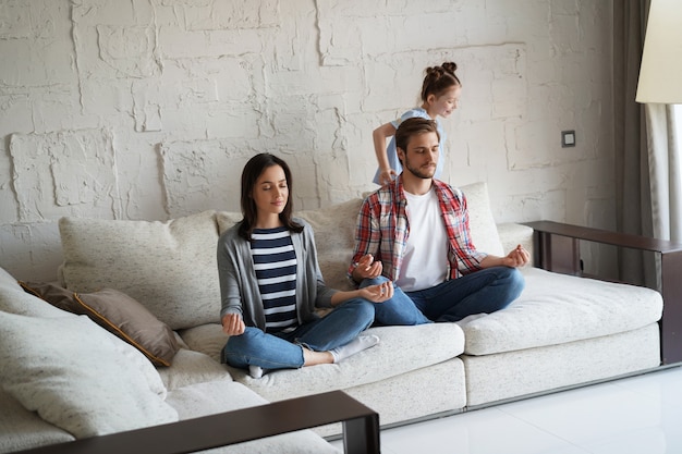 Erschöpfte Mutter und Vater, die auf der Couch sitzen, fühlen sich genervt, müde, während die laute kleine Tochter schreiend auf dem Sofa herumläuft.