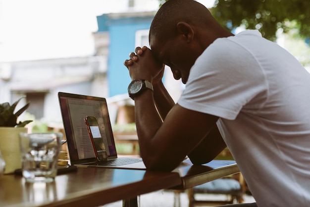 Erschöpfter, gestresster Mann mit dunkler Haut, der seinen Kopf auf gemeinsame Hände in der Nähe von Laptop und Smartphone lehnt Online-Konferenz im Café