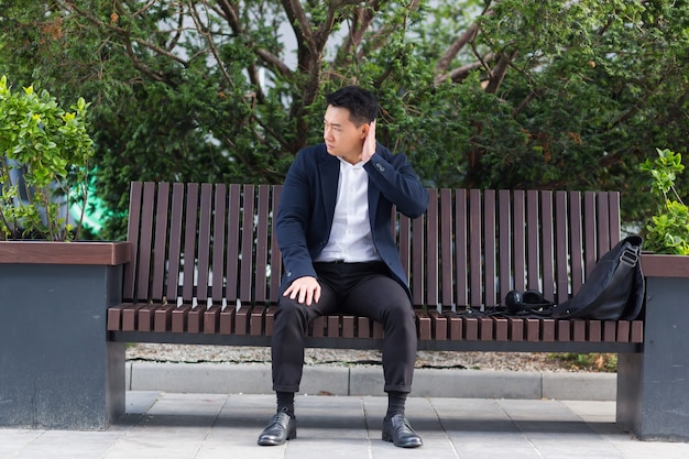Erschöpfter junger asiatischer Geschäftsmann, der draußen auf der Bank im Stadtpark sitzt. gestresster überarbeiteter Mann mit Kopfschmerzen massiert den Kopf. Depressiv Müder Mitarbeiter mit schmerzkranker Arbeit