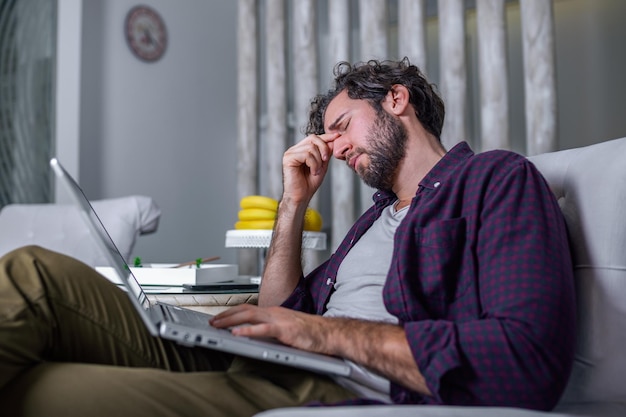 Erschöpfter müder Geschäftsmann, der zu Hause am Laptop arbeitet, Nasenrücken massiert, Müdigkeitsbeschwerden verspürt, gut aussehender junger Mann, der zu Hause unter Kopfschmerzen leidet, Nahaufnahme