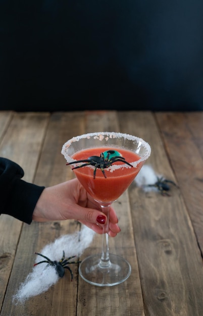 Erschreckender roter Cocktail mit Spinne auf schwarzem Hintergrund und Halloween-Holztisch mit Dekoration vertikal