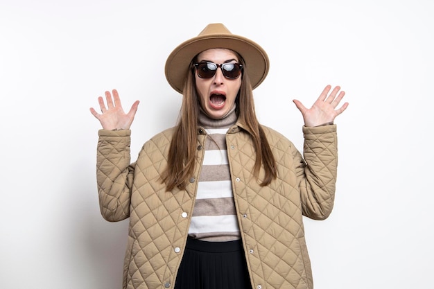 Erschrocken überraschte junge Frau in einer Jacke mit Hut gegen eine weiße Wand