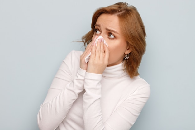 Erschrockene junge Frau im weißen Rollkragenpullover mit der Serviettenblasnase, die auf die Quelle der Allergie schaut.