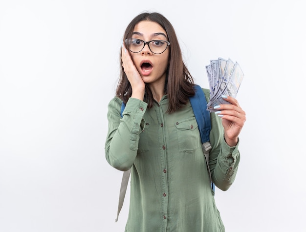 Erschrockene junge Schulfrau mit Brille mit Rucksack, die Bargeld hält und Hand auf die Wange legt