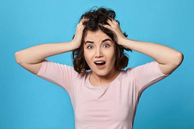 Erschrockene lockige junge Brunettefrau auf blauem Hintergrund. Menschliche Emotionen, Gesichtsausdruckkonzept.