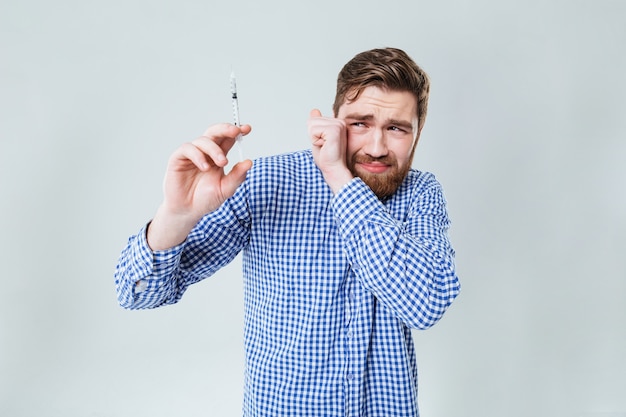 Erschrockener bärtiger junger Mann mit Spritze über weißer Wand