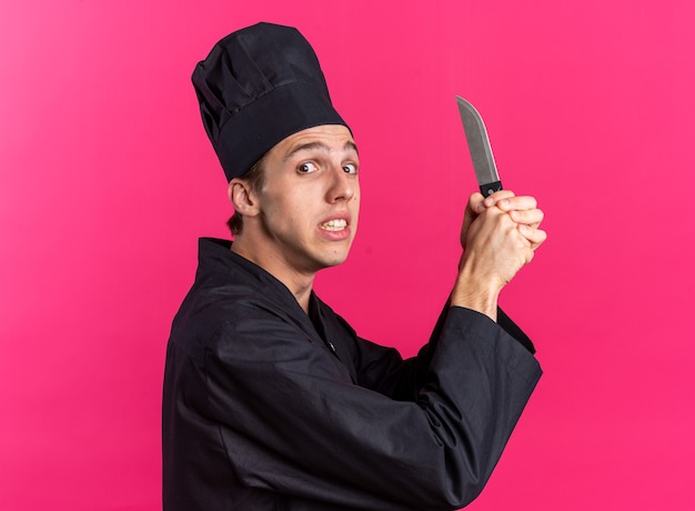 Erschrockener junger blonder männlicher Koch in Kochuniform und Mütze, der in der Profilansicht steht und Messer mit beiden Händen hält, die isoliert auf die rosa Wand blicken?