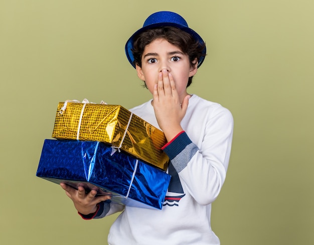 Erschrockener kleiner Junge mit blauem Partyhut, der Geschenkboxen hält, bedeckte den Mund mit der Hand