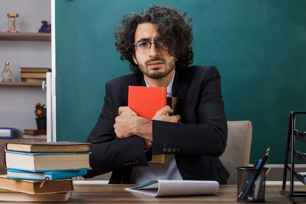 Erschrockener männlicher Lehrer mit Brille, der ein Buch hält, das am Tisch mit Schulwerkzeugen im Klassenzimmer sitzt