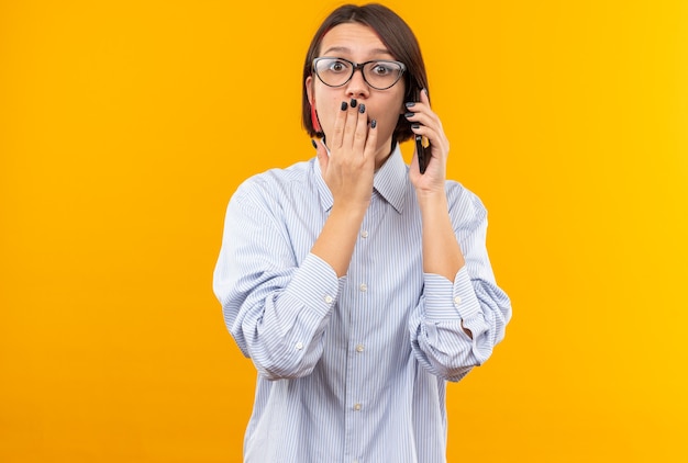 Erschrockenes junges schönes Mädchen mit Brille spricht am Telefon bedeckten Mund mit der Hand