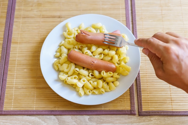 Erschwingliches Essen, eine Hand, die eine Gabel mit einer angehefteten Wurst auf dem Hintergrund eines Tellers Nudelk...