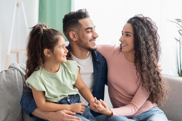 Erst die Familie. Porträt der glücklichen arabischen Mutter, des Vaters und der Tochter, die sich auf der Couch umarmen, während sie sich zusammen im Wohnzimmer entspannen, fröhliche Eltern aus dem Nahen Osten, die sich mit ihrem weiblichen Kind verbinden, Nahaufnahme