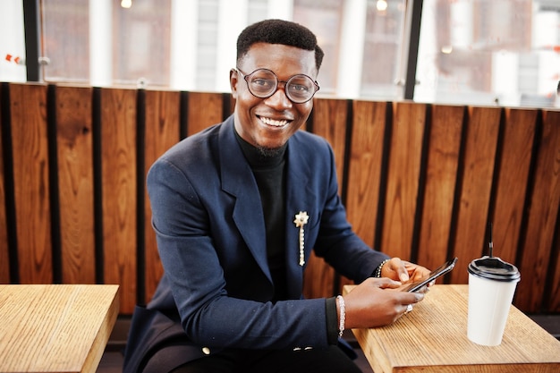 Erstaunlich aussehender afroamerikanischer Mann trägt einen blauen Blazer mit Brosche, schwarzem Rollkragenpullover und Brille, die auf der Straße gestellt werden Modischer schwarzer Typ mit einer Tasse Kaffee, der auf sein Telefon schaut