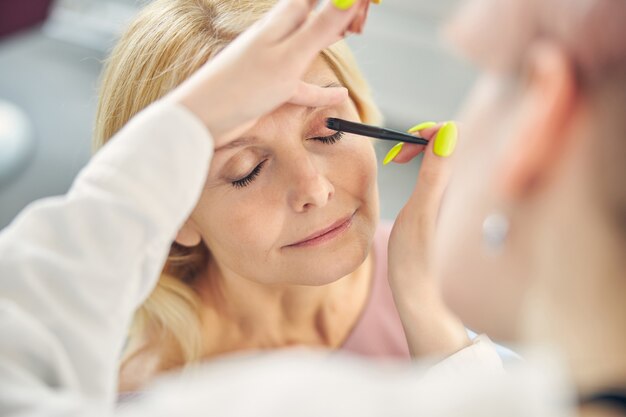 Erstaunliche blonde Frau, die Positivität ausdrückt, während sie professionelles Make-up für ein Treffen im Salon macht