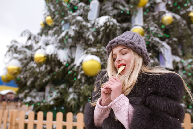 Erstaunliche blonde Frau in grauer Strickmütze beißt köstliche Pralinen gegen Weihnachtsfichte mit Lichtern