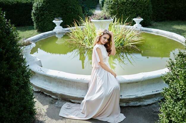 Erstaunliche Braut mit dem langen lockigen Haar im Hochzeitskleid, das nahe Brunnen am Parkhintergrund, Hochzeitsfoto, Porträt sitzt.