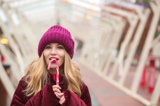 Erstaunliche junge blonde Frau in roter Strickmütze posiert mit Zuckerstange auf der Straße