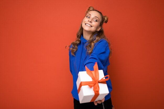 erstaunliche junge blonde Frau lokalisiert über rote Hintergrundwand, die blauen stilvollen Hoodie hält, der Geschenkbox hält und Kamera betrachtet.
