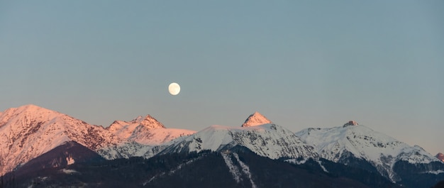 Erstaunlicher Sonnenuntergang und Vollmond über den schneebedeckten Bergen des Kaukasus in Krasnaya Polyana, Russland.