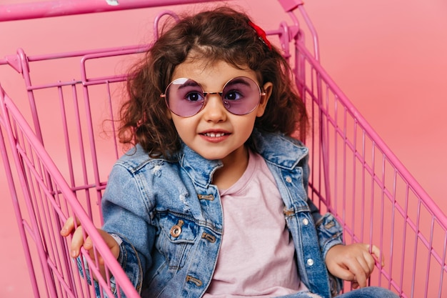 Erstaunliches lockiges Kind, das im Einkaufswagen sitzt Studioaufnahme eines Mädchens im Vorschulalter mit Sonnenbrille