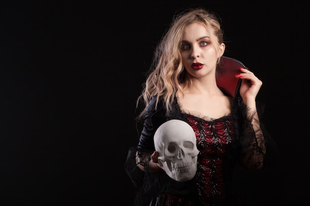 Erstaunliches totes Mädchen, das wie ein Vampir verkleidet ist, der einen Schädel für Halloween-Karneval hält. Porträt der Frau im Vampirkostüm.