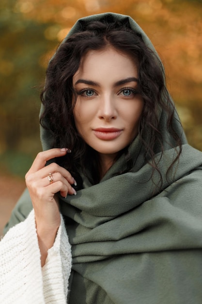 Erstaunliches weibliches Herbstporträt einer schönen jungen Frau mit lockigem Haar und magischen blauen Augen in einem modischen Strickpullover mit einem grünen Schal auf dem Kopf, der in einem Herbstpark spazieren geht