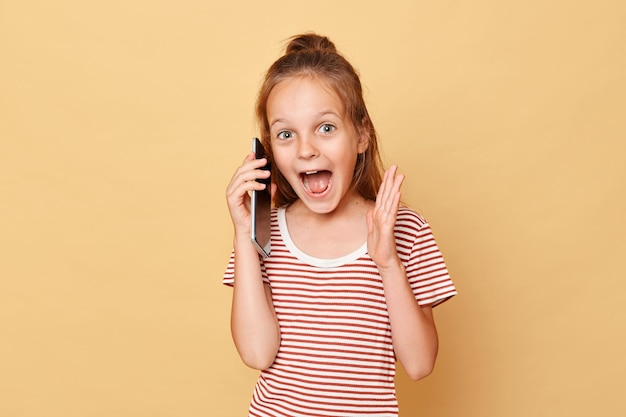 Erstaunt, aufgeregt, kleines braunhaariges Mädchen in gestreiftem T-Shirt, das isoliert vor beigem Hintergrund steht und schreiend mit dem Handy spricht und hervorragende Nachrichten hört