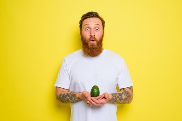 Erstaunt mit Bart und Tattoos hält eine Avocado