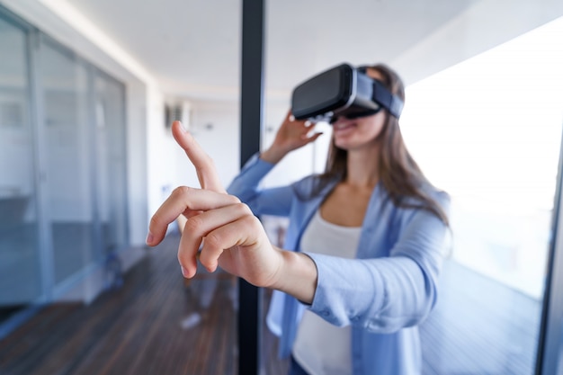 Erstaunt überraschte junge Frau mit Virtual-Reality-Brille, die während des VR-Erlebnisses zu Hause die Luft berührt