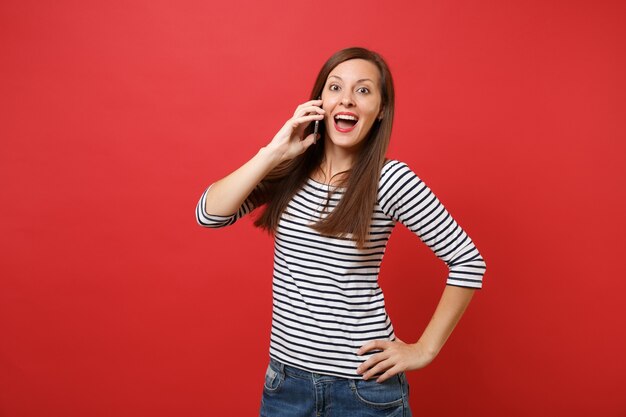 Erstaunte Frau, die den Mund weit offen hält, überrascht aussieht, mit dem Handy spricht, angenehme Gespräche auf rotem Hintergrund führt. Menschen aufrichtige Emotionen, Lebensstil. Kopieren Sie Platz.