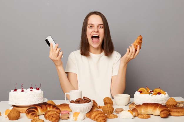 Erstaunte Frau mit braunen Haaren, die mit Handy in der Hand zwischen verschiedenen Desserts am Tisch sitzt, hat guten Appetit, aufgeregt mit einer Vielzahl von Süßwaren, die müde von Diäten sind, die auf grauem Hintergrund isoliert sind