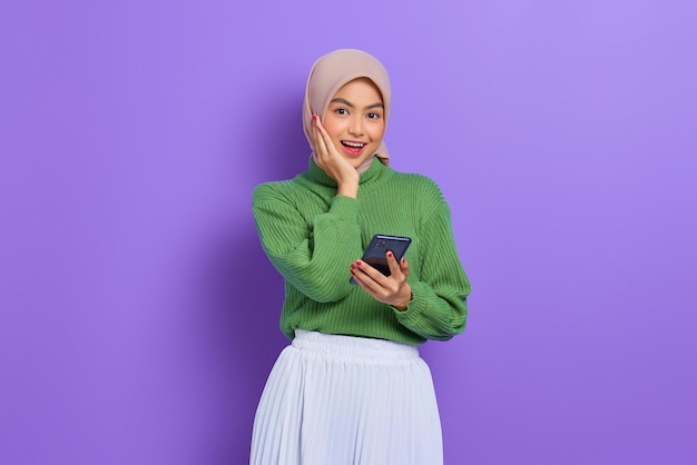 Erstaunte fröhliche schöne asiatische Frau in grünem Pullover und Hijab mit Handy isoliert über violettem Hintergrund