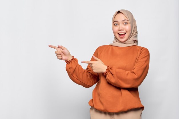 Erstaunte junge Asiatin im orangefarbenen Hemd, die mit dem Finger auf den Kopierraum zeigt, der über weißem Hintergrund isoliert ist