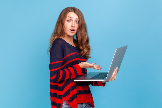 Erstaunte junge Frau, die einen gestreiften Pullover im lässigen Stil trägt, Laptop in den Händen hält, schockiert über Arbeitsanforderungen, Frist. Studioaufnahme im Innenbereich isoliert auf blauem Hintergrund.