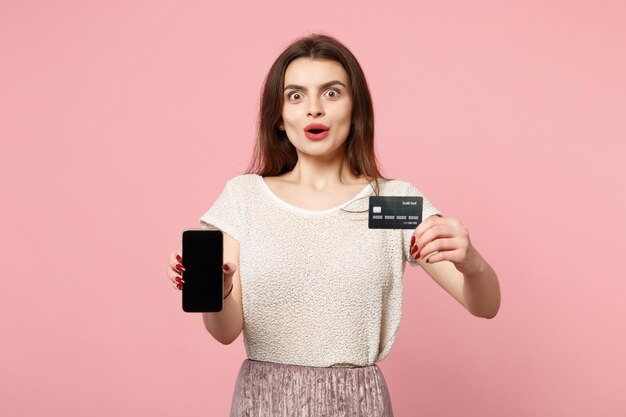 Erstaunte junge Frau in lässiger leichter Kleidung, die isoliert auf pastellrosa Wandhintergrund posiert. People-Lifestyle-Konzept. Mock-up-Kopienbereich. Holding-Handy mit leerem, leerem Bildschirm, Kreditkarte.