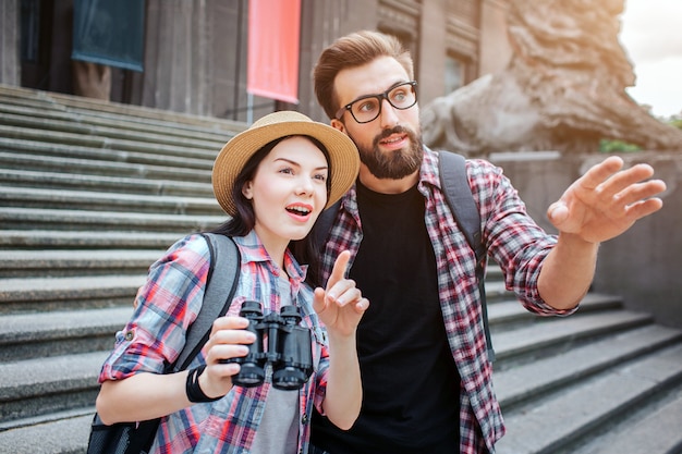 Erstaunte Touristen stehen vor Treppen. Sie zeigen nach vorne. Frau hält Fernglas. Sie sind in der Stadt. Bärtiger Mann trägt eine Brille.