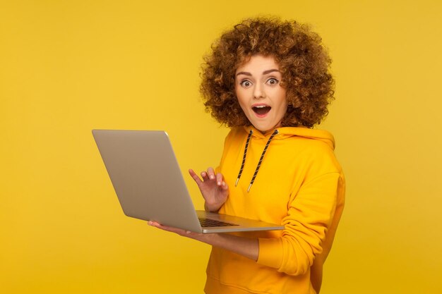 Erstaunte überraschte Frau mit Afro-Frisur, die einen Laptop hält und mit schockiertem Ausdruck aussieht, die am Computer arbeitet und einen Hoodie im lässigen Stil trägt. Studioaufnahme im Innenbereich isoliert auf gelbem Hintergrund