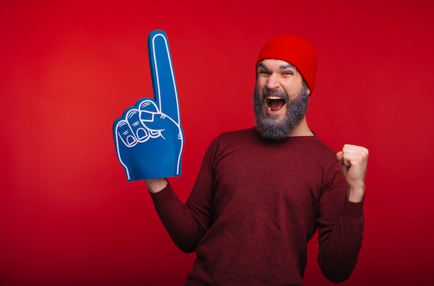 Erstaunter bärtiger Hipster-Mann, der über rotem Raum steht und Erfolg mit Fächerfingerhandschuh feiert