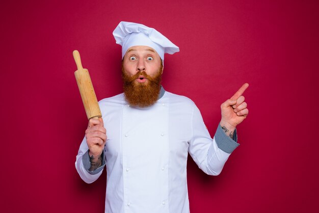 Erstaunter Koch mit Bart und roter Schürze Koch hält hölzernes Nudelholz